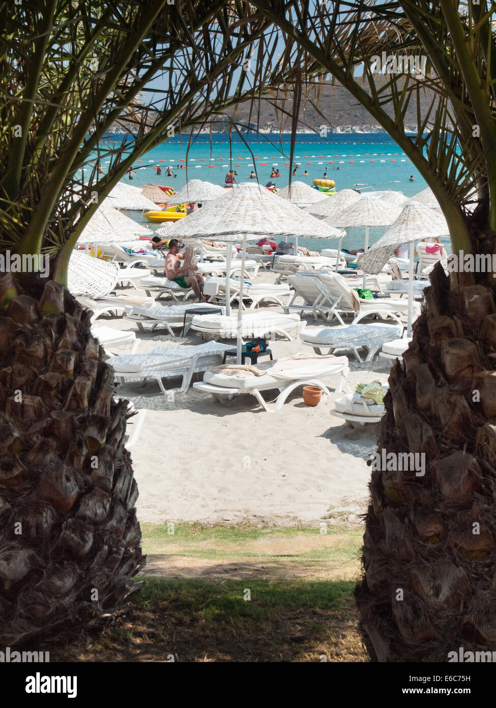 Cesme, Turkey,  August 2nd, 2014: Tourist beach on the Cesme peninsula in high season viewed through palm trees. Stock Photo