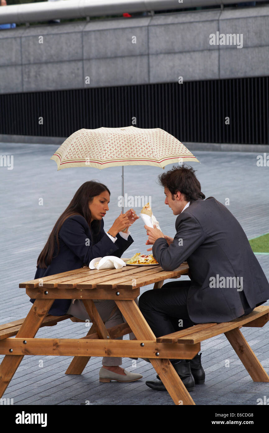 Young couple sitting on bench eating lunch sheltering under umbrella in the rain at Southbank, London in August Stock Photo