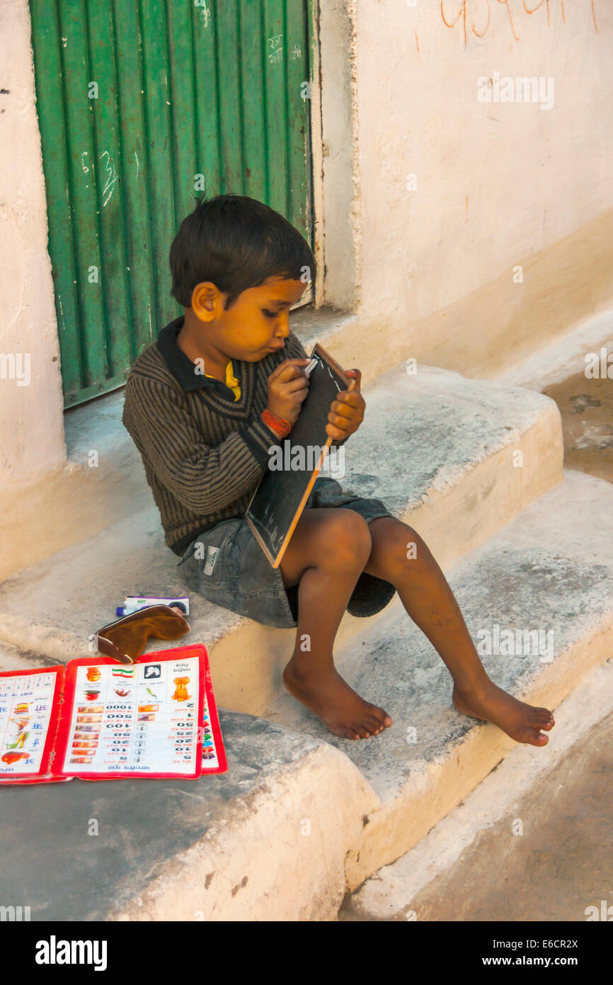 Barefoot kid on the steps of his public school scratching with chalk upon a small blackboard. Stock Photo