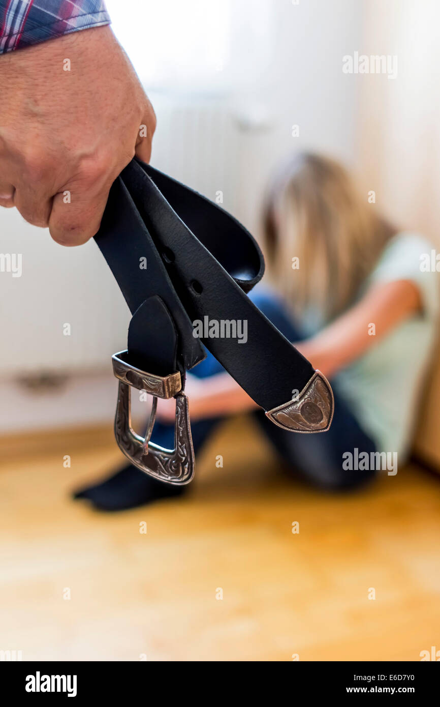 Man's hand holding belt while young woman crouching afraid on the floor Stock Photo