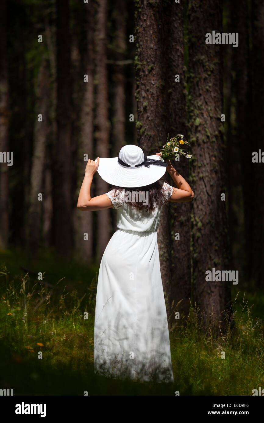 Woman from behind in white dress and white hat walking in the wood holding a bunch of flowers Stock Photo