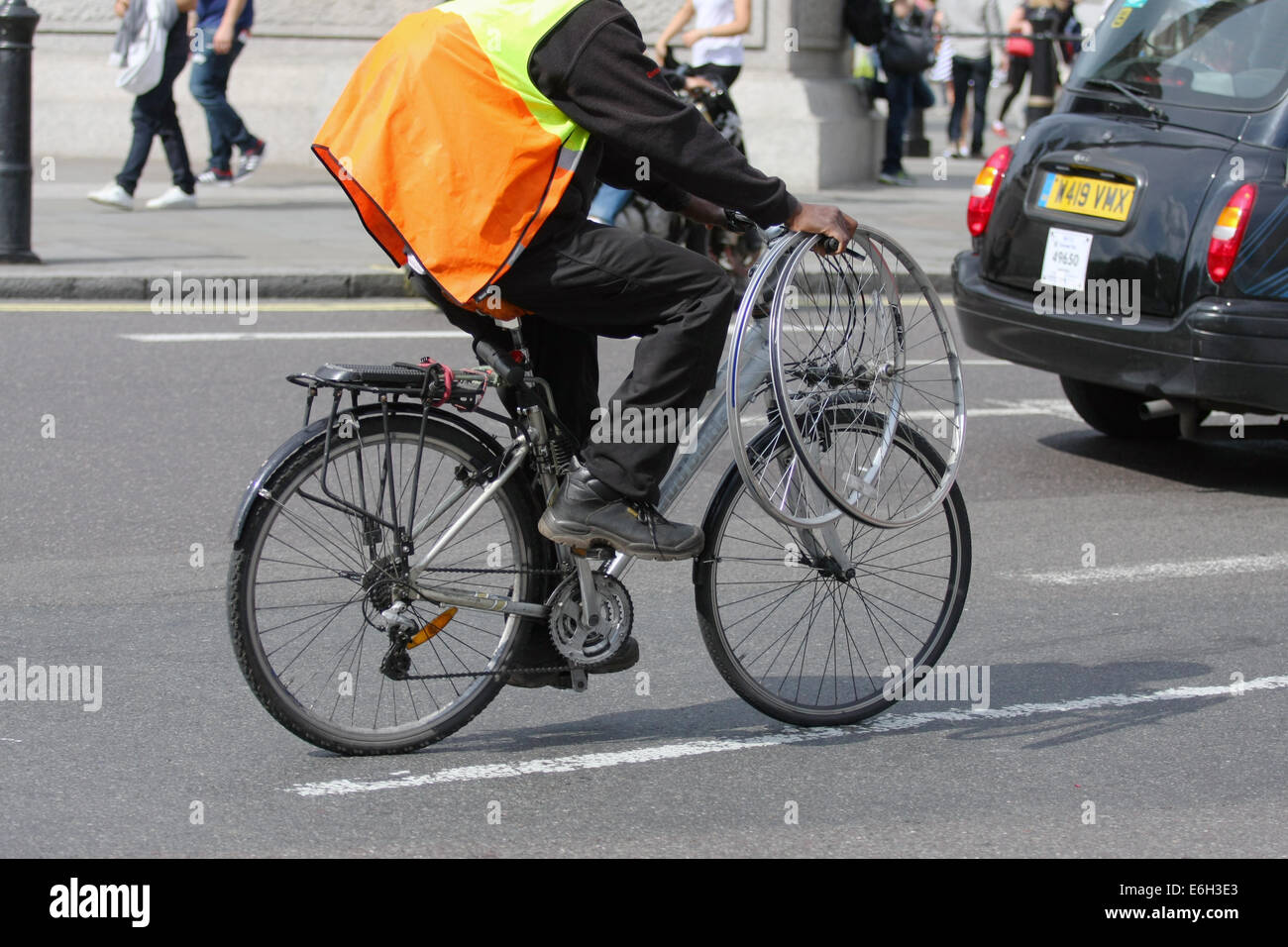 A cyclist riding a bicycle and carrying two spare wheels behind a taxi, in London. Stock Photo