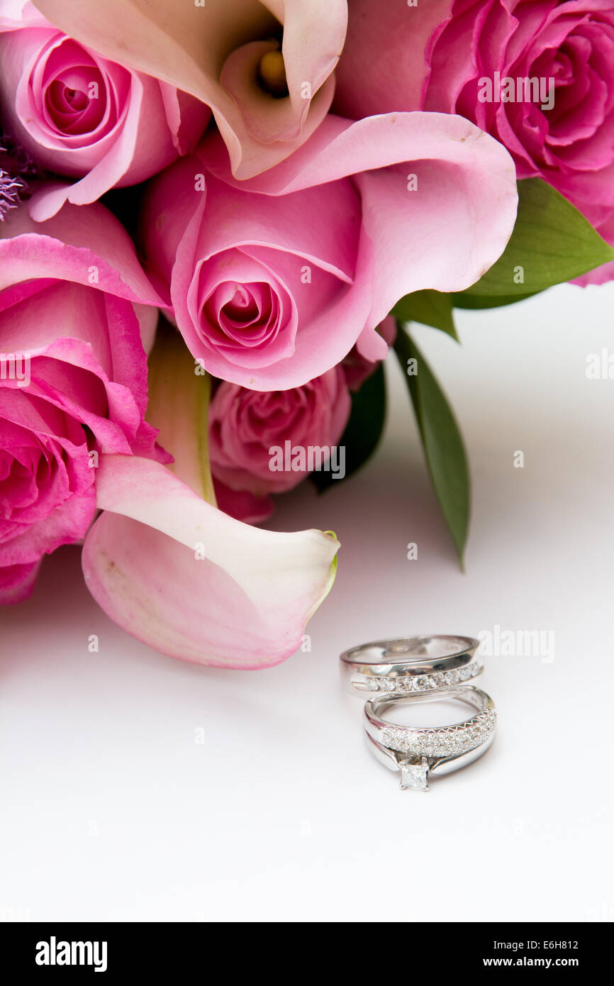White gold and diamond wedding rings and an engagement ring are displayed on a white background next to pink wedding flowers. Stock Photo