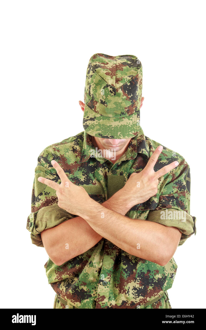 unknown soldier with hidden face in green camouflage uniform covers face with cap and standing with sign of peace with cross arm Stock Photo