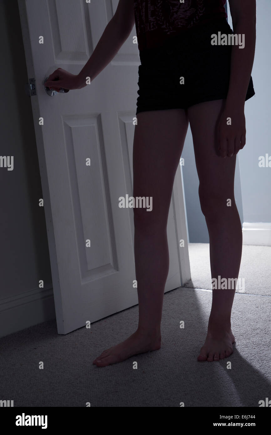 Young female standing in a darkened room with her hand on the doorknob, shadows cast by light from behind. Lower body shot. Stock Photo