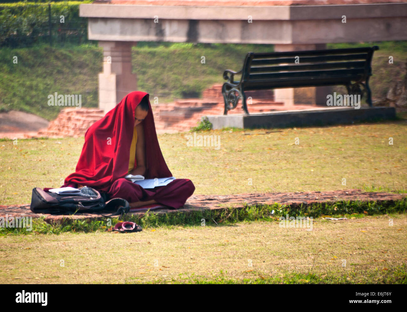 Young monk with his head partly and his body covered by a red sheet of cloth, sitting cross legged at Dhamekh Stupa. Stock Photo