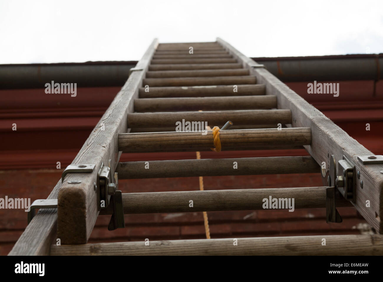 A very tall wooden extension ladder reaches up to the roof of a brick building.  Extension locks are visible. Stock Photo