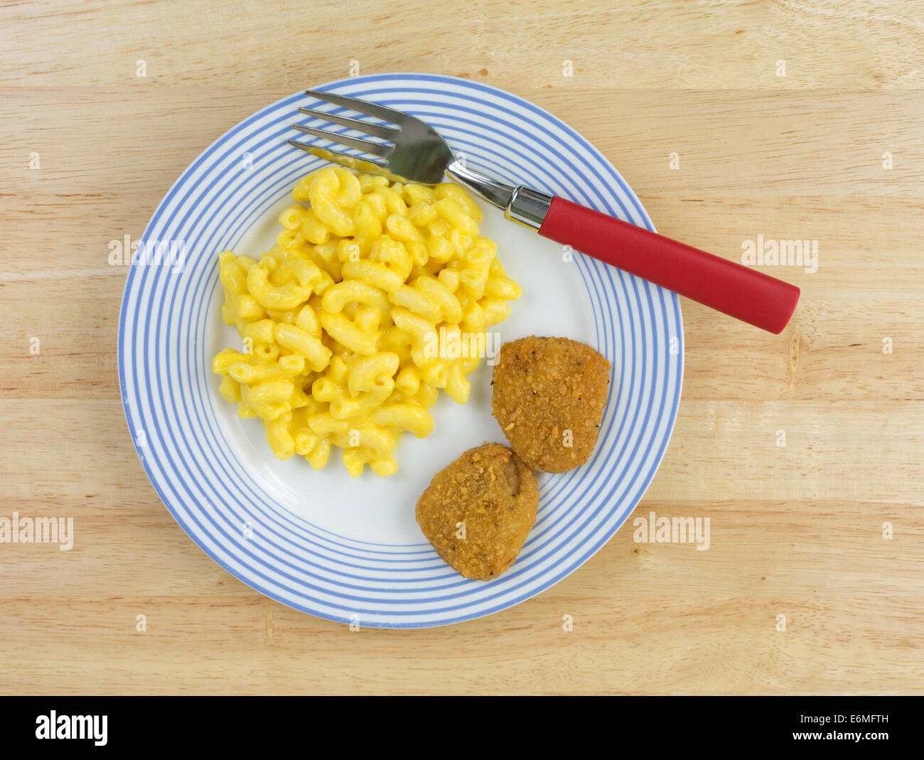 Top view of a meal of macaroni and cheese with two chicken nuggets on a blue striped plate with a red handled fork on a wood tab Stock Photo