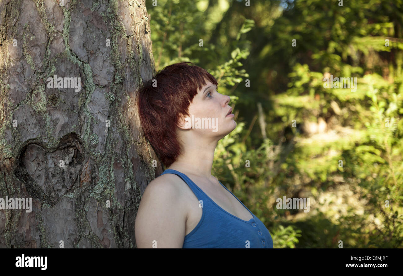 Beautiful redhead girl leaning on a tree in a forest and daydreaming. Tree has a heart engraving. Stock Photo
