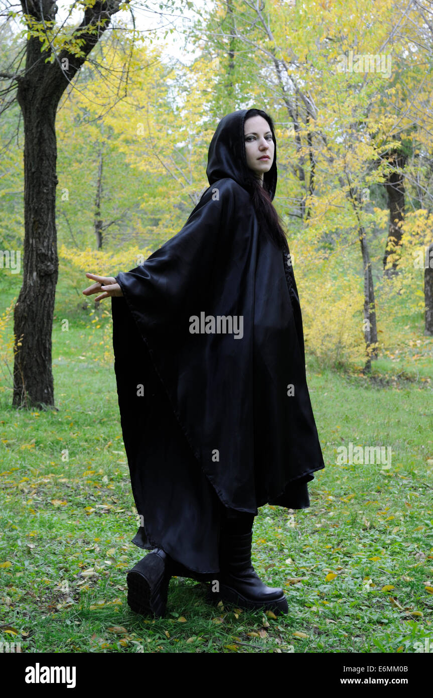 Black hooded dryad woman in the autumn forest, turning back at you while walking as if beckoning Stock Photo