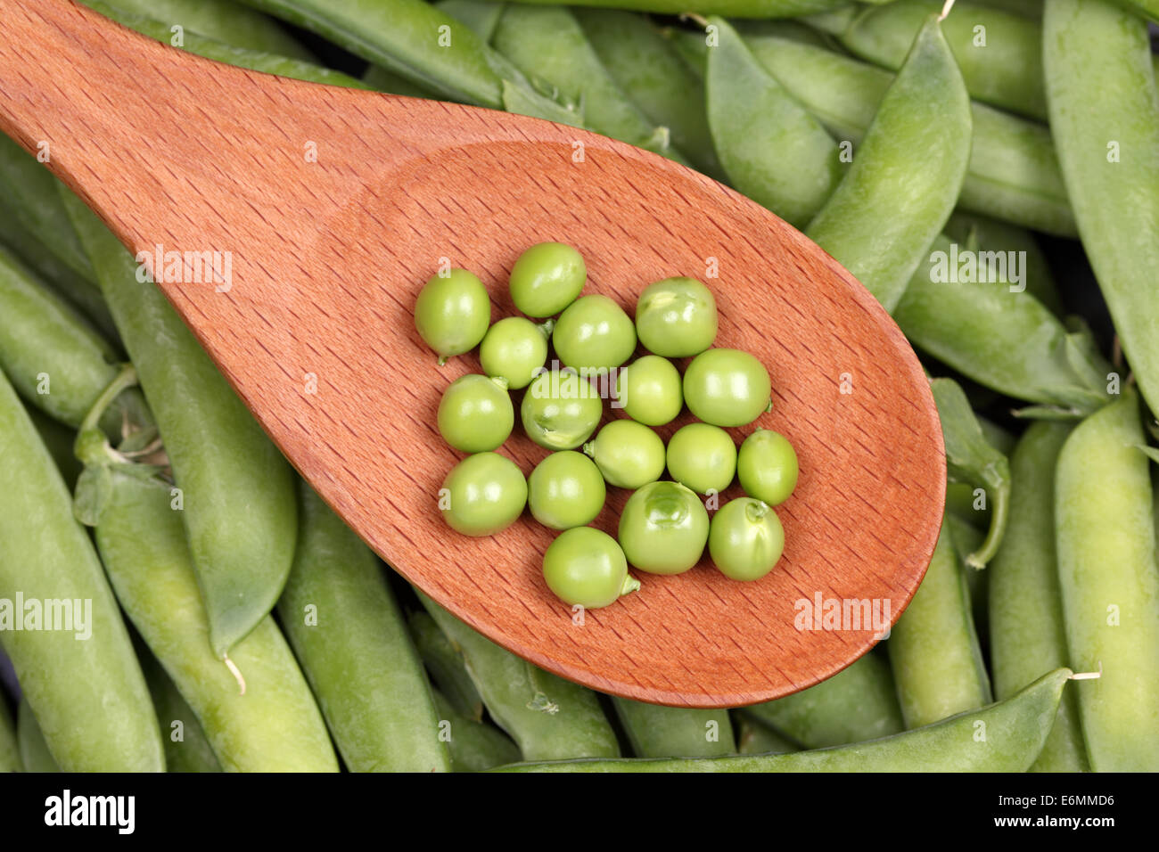 Green peas in a wooden spoon on green pea in the pod background. Stock Photo