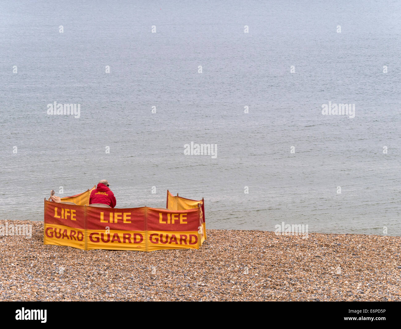 Lone seaside lifeguard behind windbreak on beach watches empty sea on cold August day, Brighton, East Sussex, England, UK Stock Photo