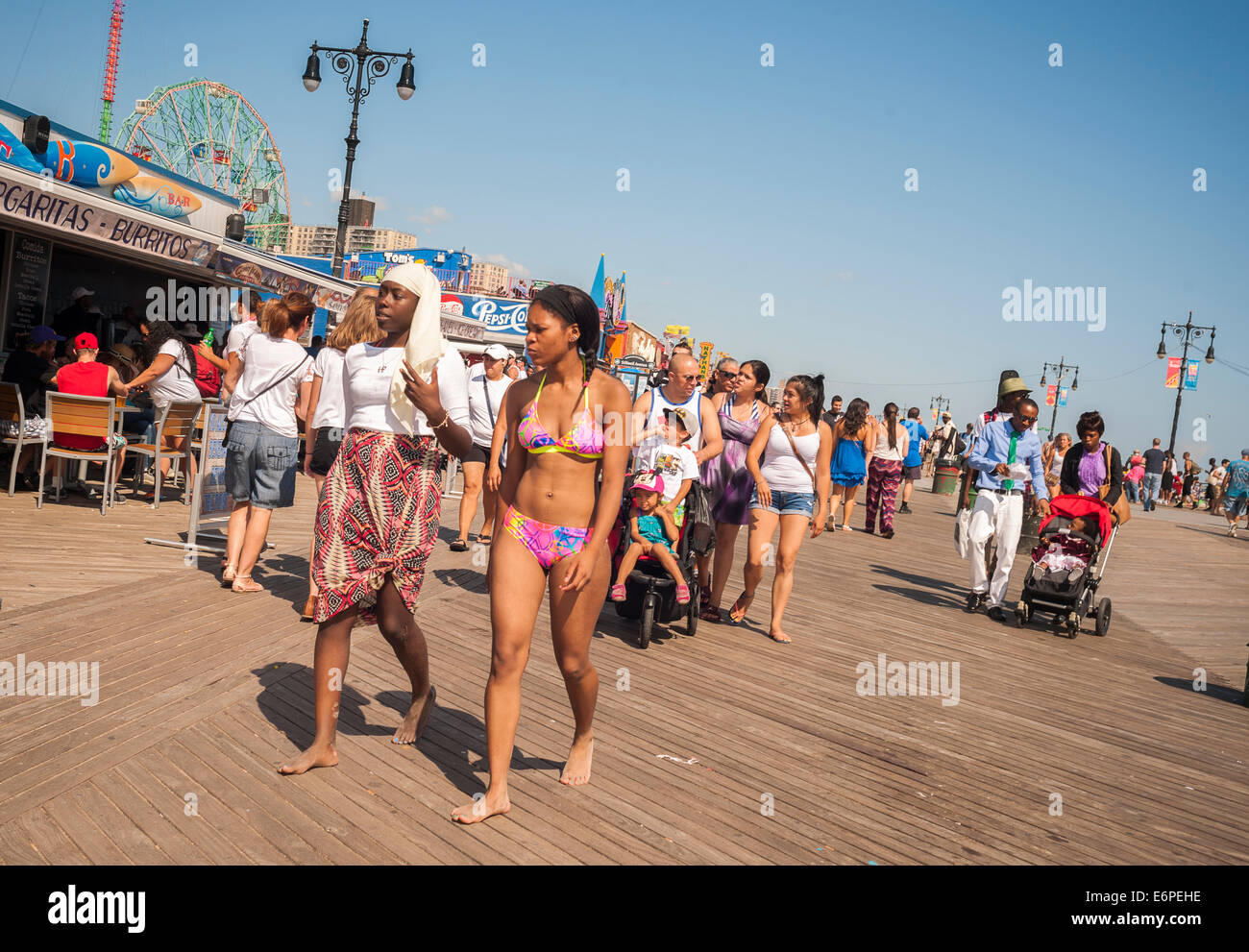 Young women, one in traditional clothing and the other in a bikini, join the crowds on the boardwalk at Coney Island in Brooklyn Stock Photo
