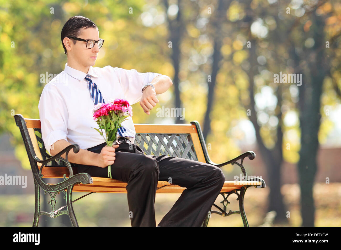 Young man holding flowers and checking the time seated on bench in park ...