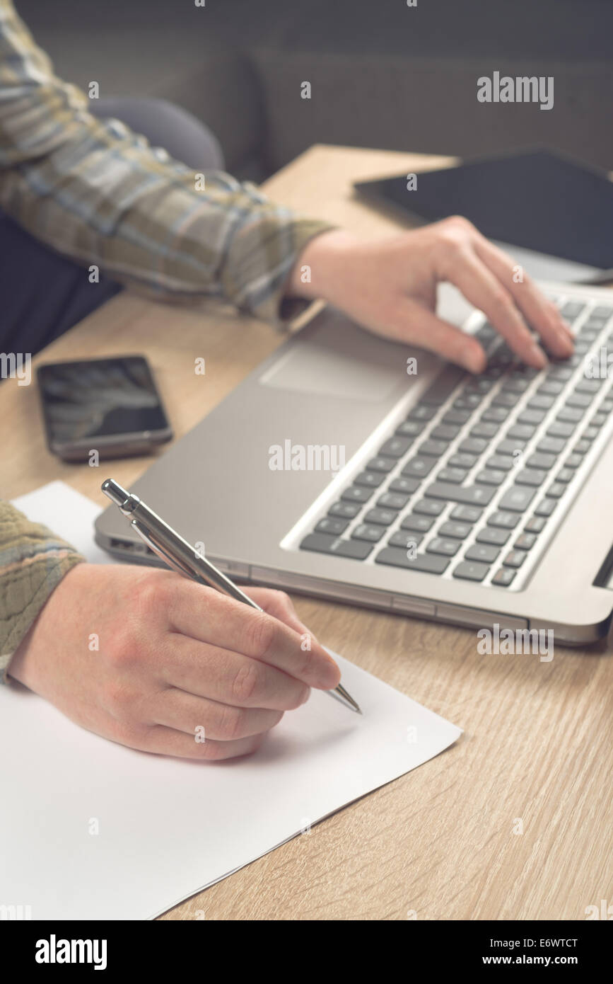 Female programmer writing notes on piece of paper while typing laptop keyboard, side view. Stock Photo