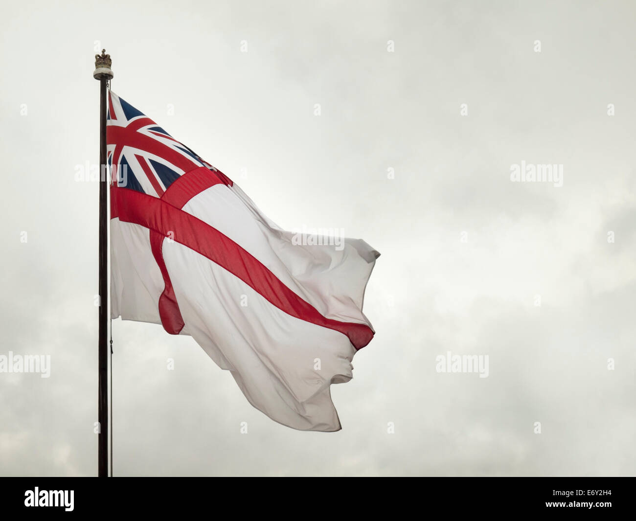 the white ensign of the British royal navy flying in a strong wind with gray overcast sky behind Stock Photo