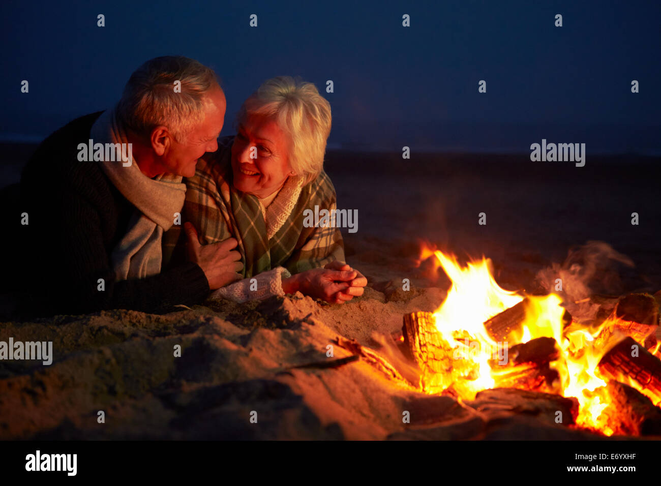 Senior Couple Sitting By Fire On Winter Beach Stock Photo