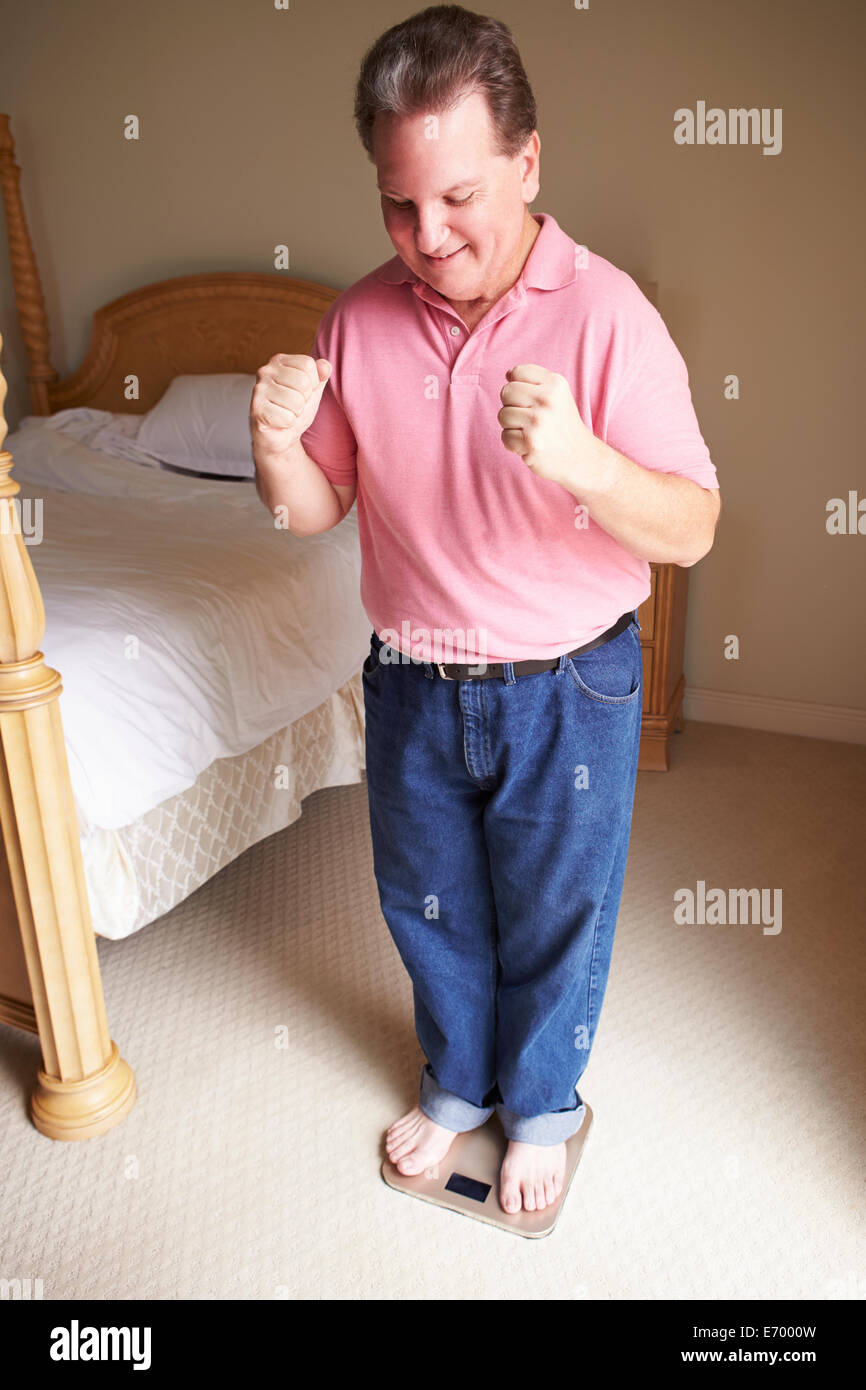 Happy Overweight Man Standing On Scales In Bedroom Stock Photo