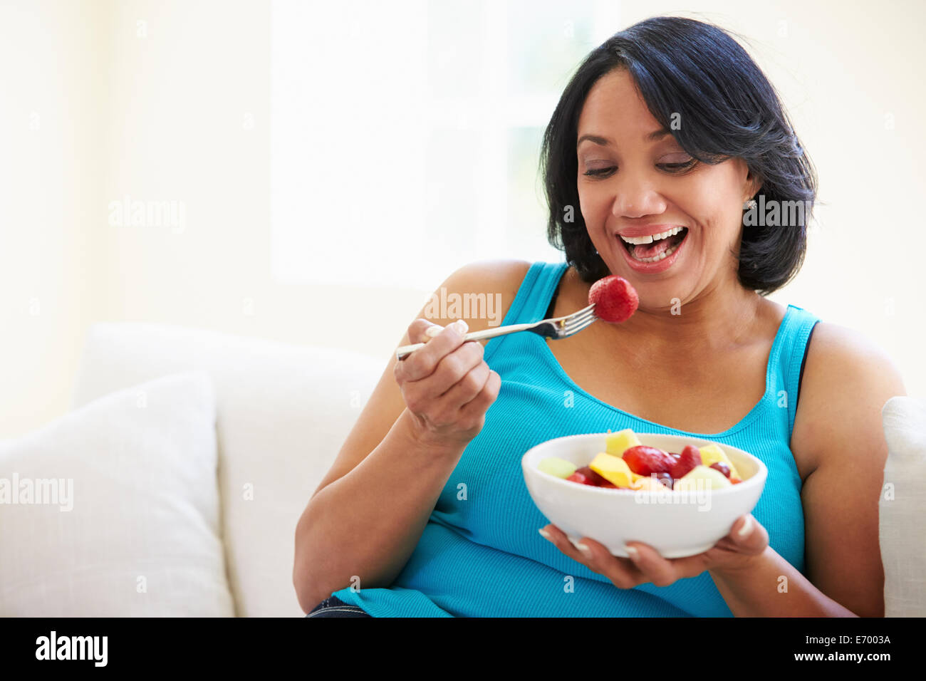 Overweight Woman Sitting On Sofa Eating Bowl Of Fresh Fruit Stock Photo