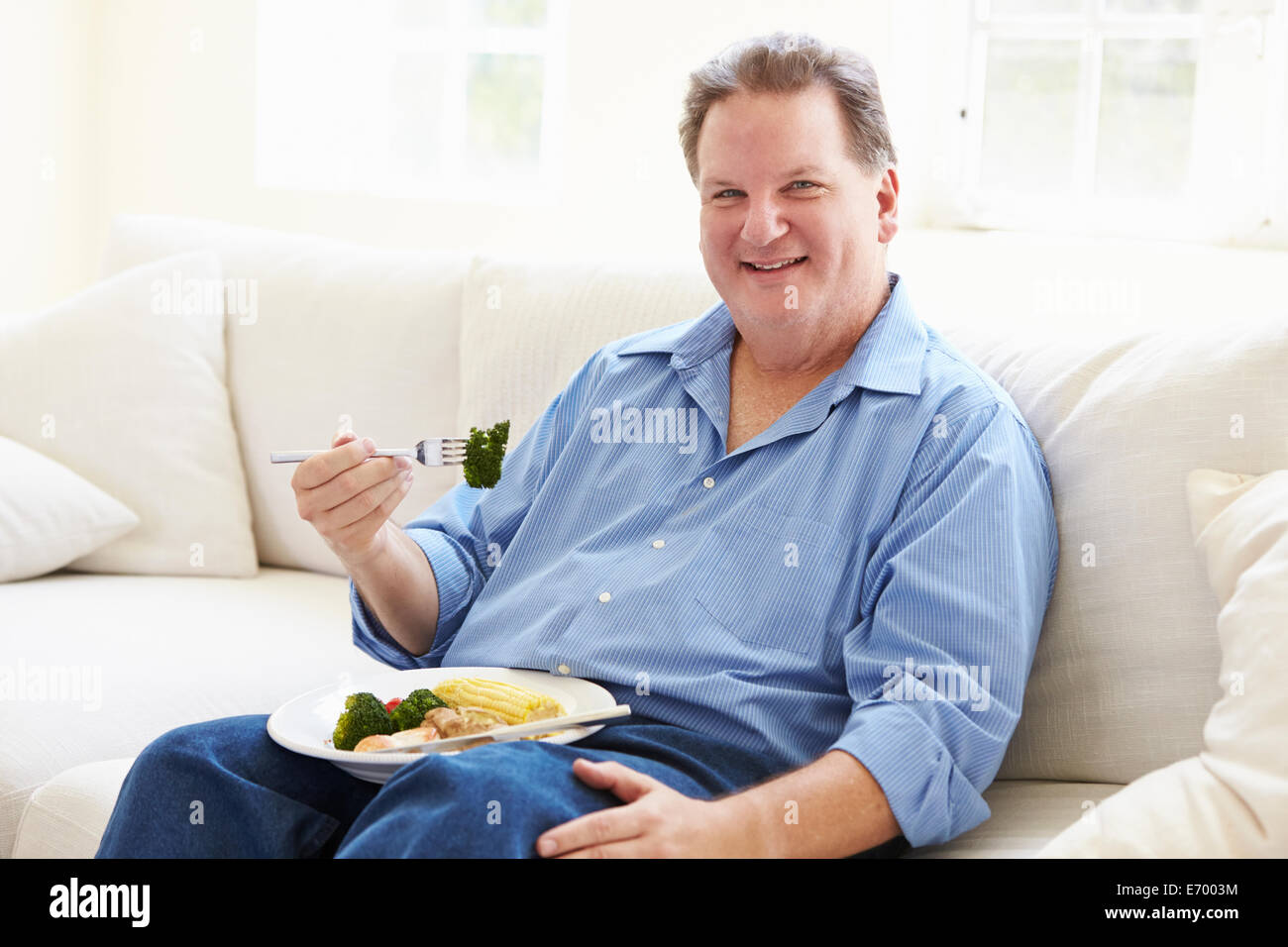 Overweight Man Eating Healthy Meal Sitting On Sofa Stock Photo