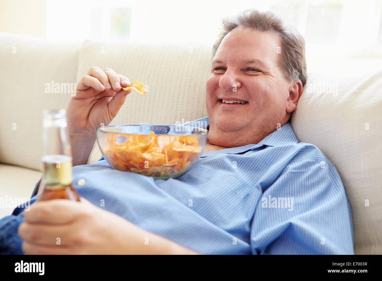 Overweight Man At Home Eating Chips And Drinking Beer Stock Photo