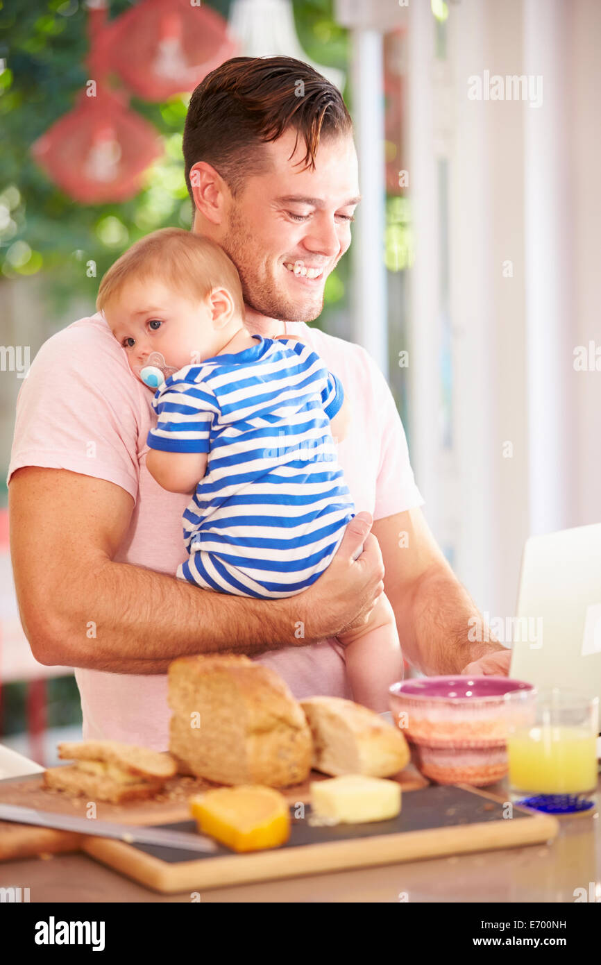 Father Holding Baby And Making Snack Whilst Using Laptop Stock Photo