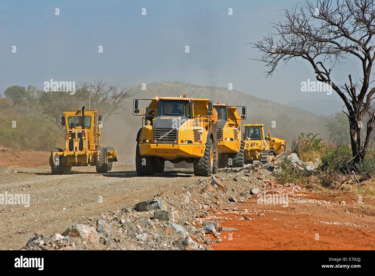 Grader vehicle working beside dump haul trucks which wait in turn to tip rocks during construction of access road to new platinum mine. South Africa Stock Photo