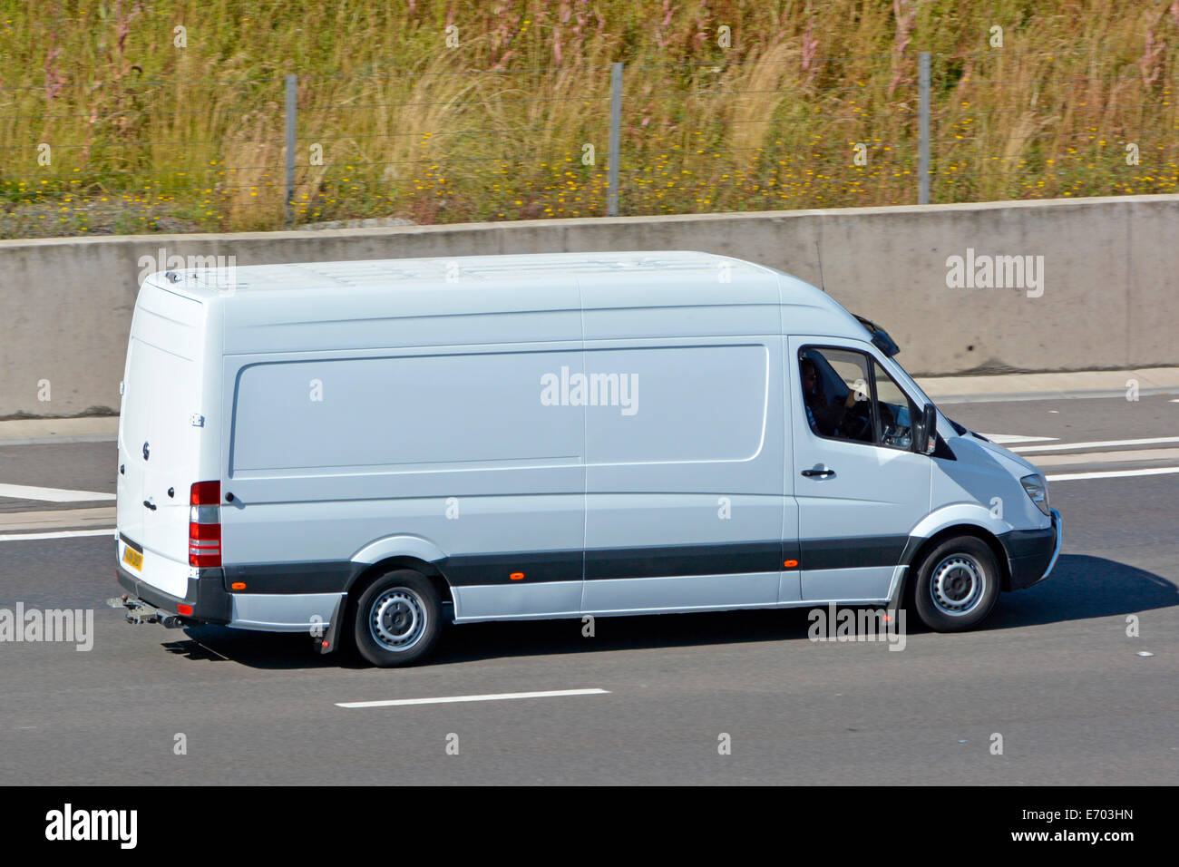 Close up side and aerial view of white clean unmarked van and driver  driving along UK motorway grass embankment and concrete retaining wall beyond Stock Photo