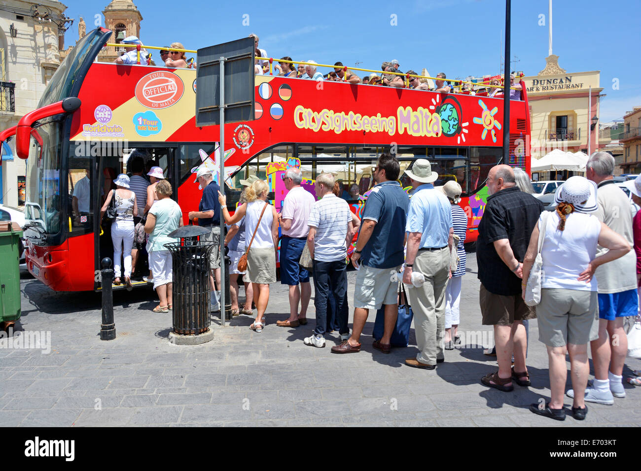 Back view line of tourists boarding & queuing up on a hot summers day to board open top Malta sightseeing tour bus at fishing village of Marsaxlokk Stock Photo