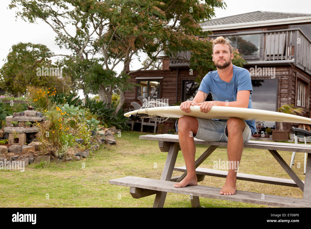 Portrait of male surfer sitting on picnic bench with surfboard on lap Stock Photo