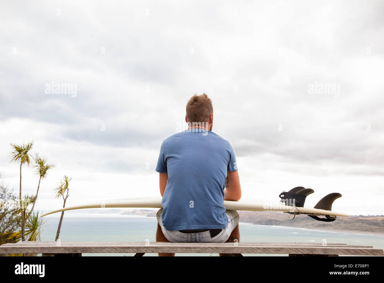 Young male surfer sitting on picnic bench looking away to sea Stock Photo