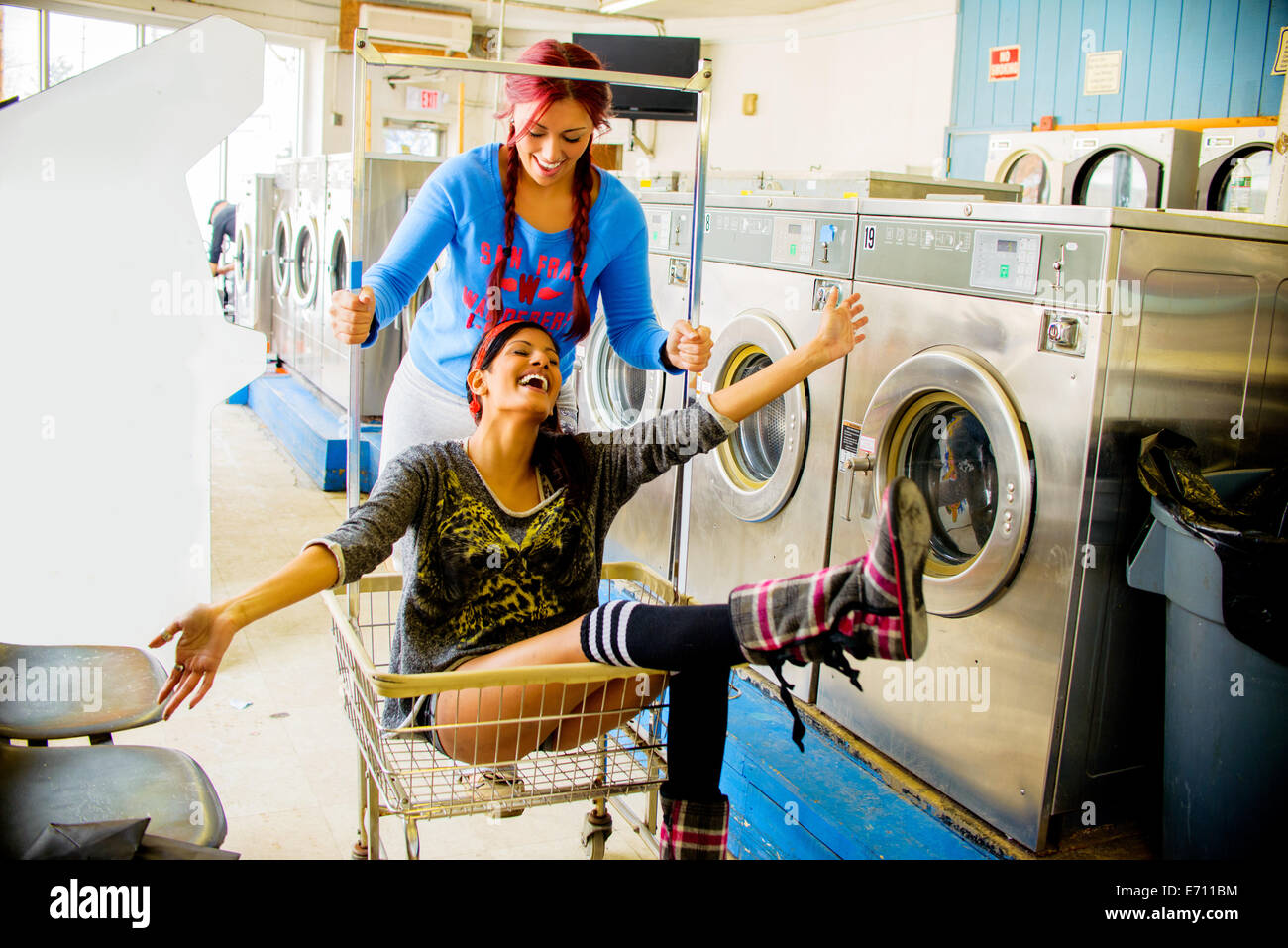 Two young women in laundromat, one pushing the other along in trolley Stock Photo