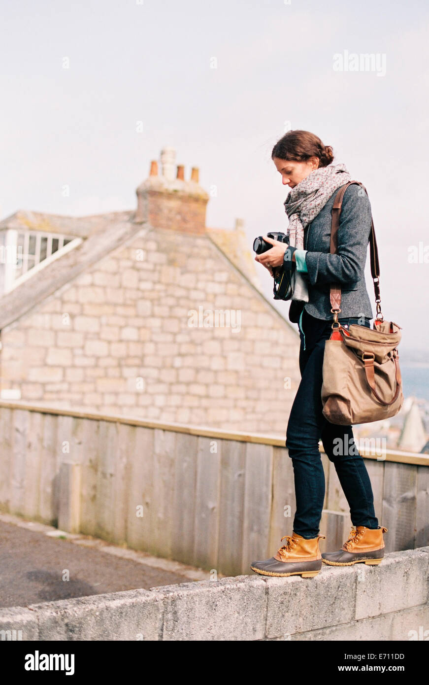A woman with a large bag, holding a camera, standing on the top of a wall. Stock Photo