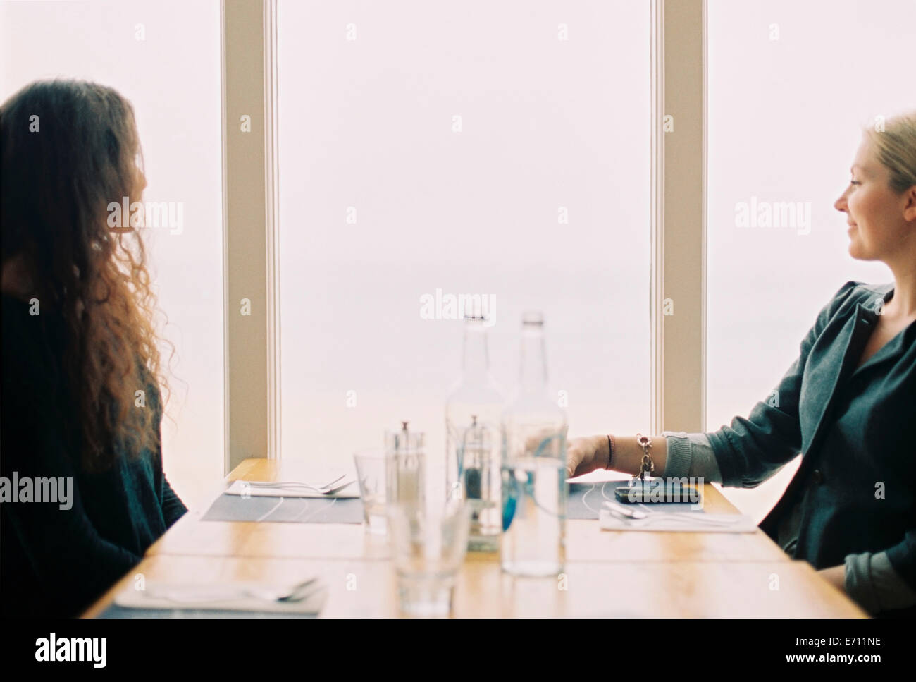 Two women seated at a table, looking out of a large window. Stock Photo