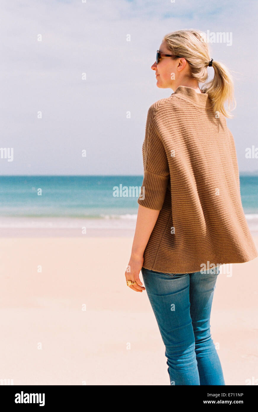 A woman in a brown jacket standing on the beach looking out to sea. Stock Photo