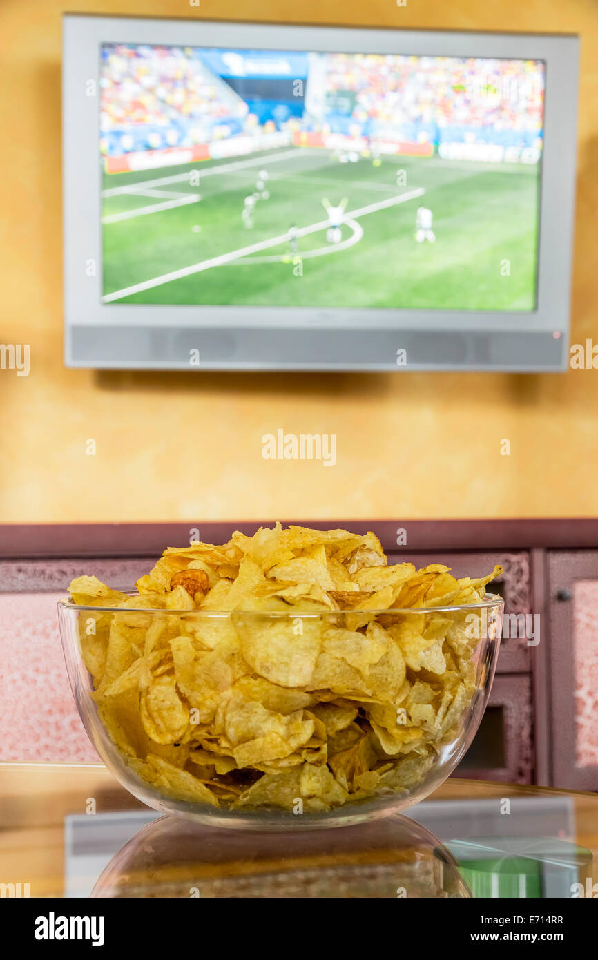 Glass bowl of potato chips in front of flatscreen TV broadcasting a soccer match Stock Photo