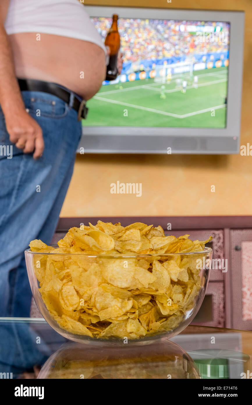 Man with naked beer belly holding beer bottle in front of flatscreen TV broadcasting a soccer match, partial view Stock Photo