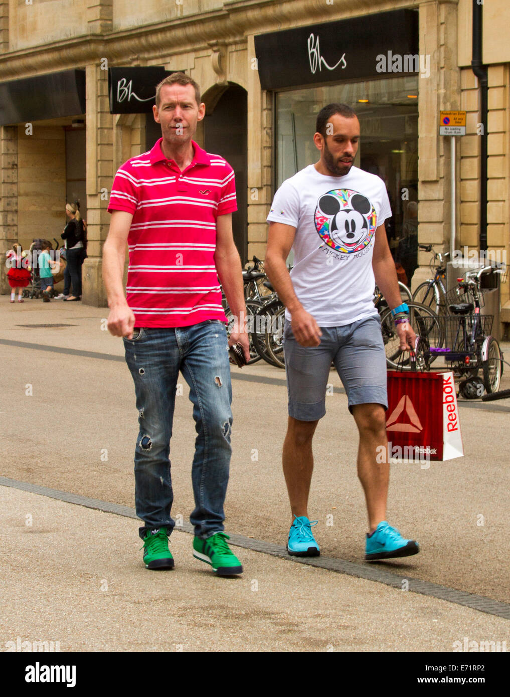 Two young men of different racial origins wearing casual clothes, one with beard and carrying shopping bag, walking along street Stock Photo