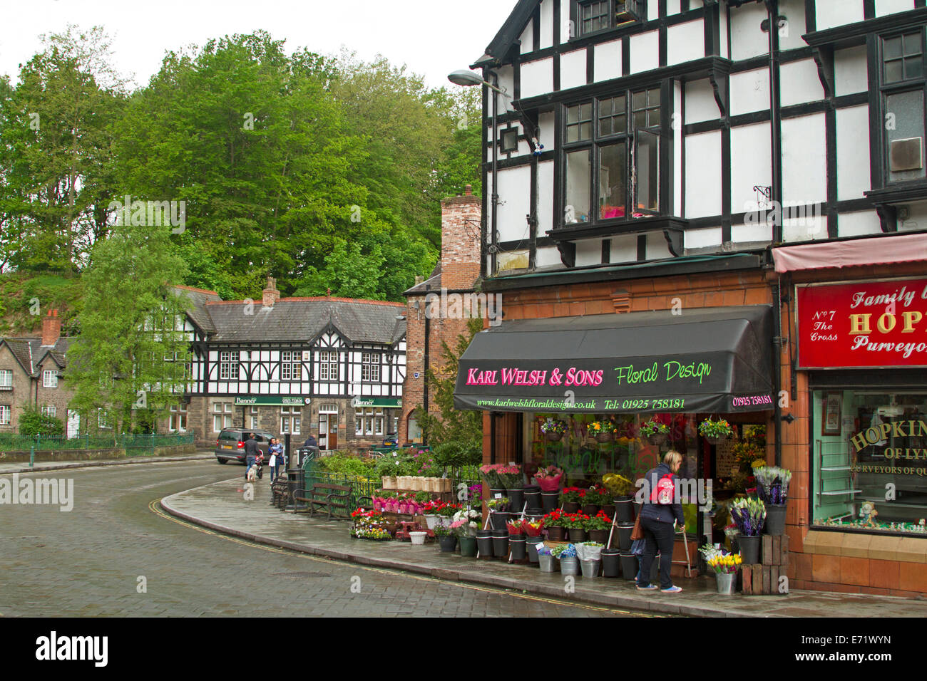 Main street and florist's shop with woman admiring colourful footpath display at picturesque village of Lymm in Cheshire, England Stock Photo