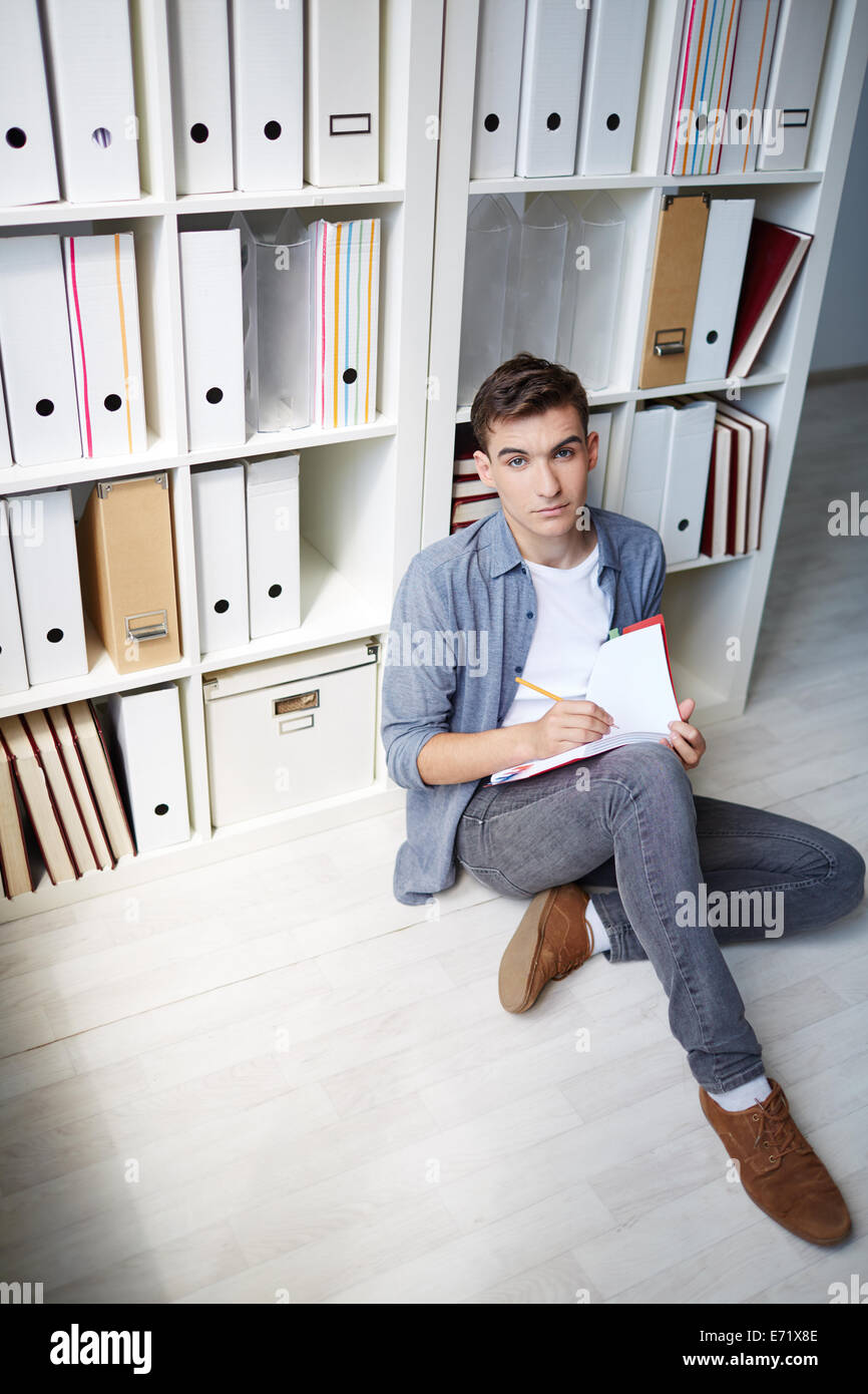 Serious student sitting on the floor and making notes in copybook Stock Photo