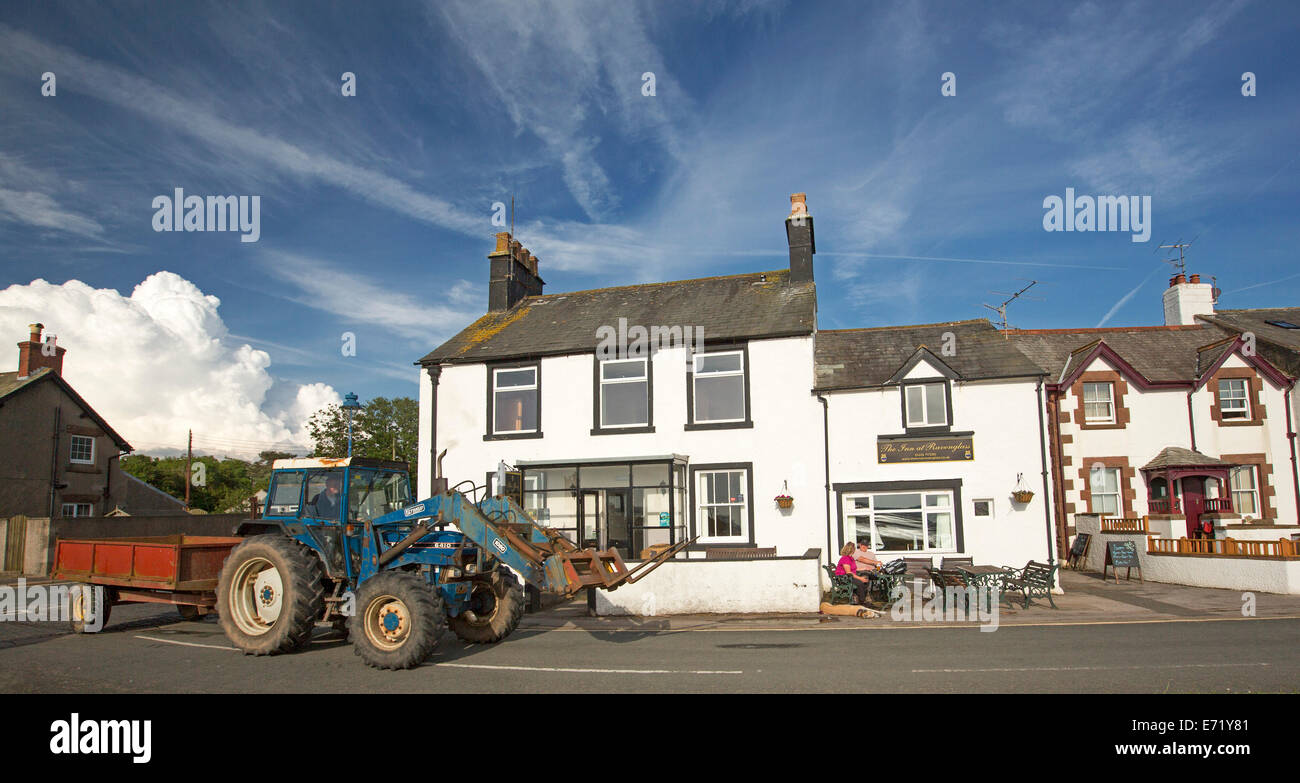Farm tractor with trailer passing beachfront houses and pub with people outside at coastal village of Ravenglass, Cumbria, England Stock Photo