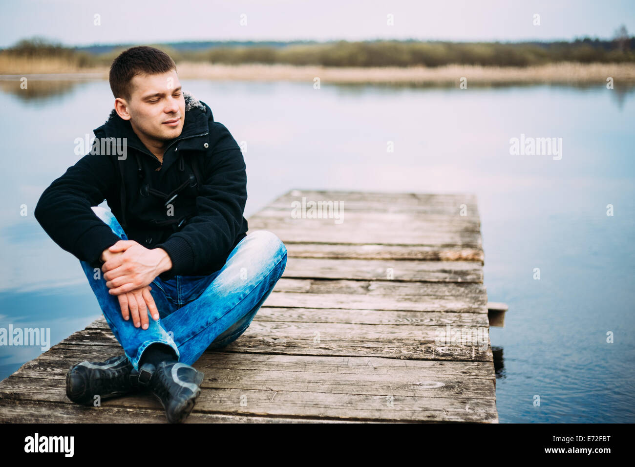 Young handsome man sitting on wooden pier in autumn day, relaxing,  thinking, listening. Casual style - jeans, jacket Stock Photo