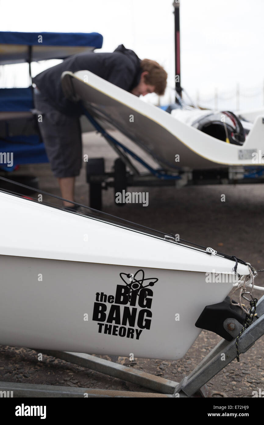A big bang theory sticker on the front of an RS300 racing sailing dinghy whilst a man works on his boat in the background. Stock Photo