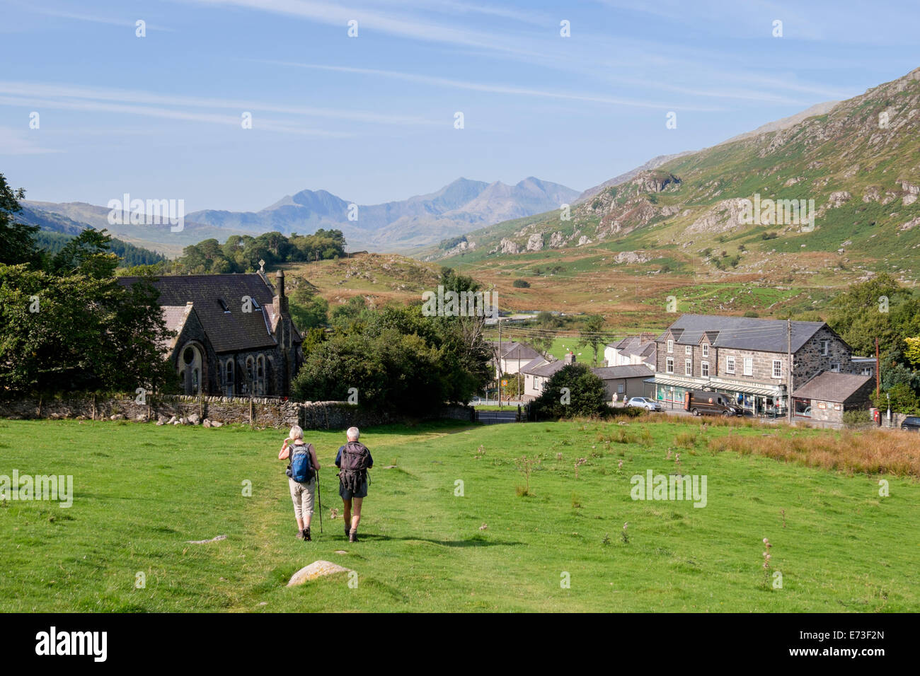 View to small village with walkers walking down hillside and distant Snowdon horseshoe mountains in late summer. Capel Curig North Wales UK Britain Stock Photo