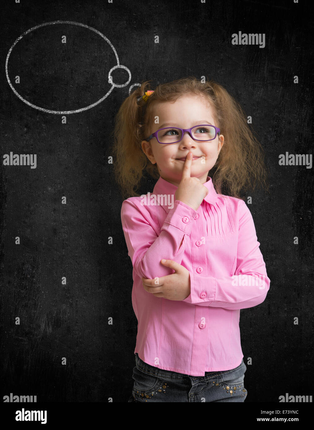 Funny child in eyeglasses standing near school chalkboard and thinking Stock Photo