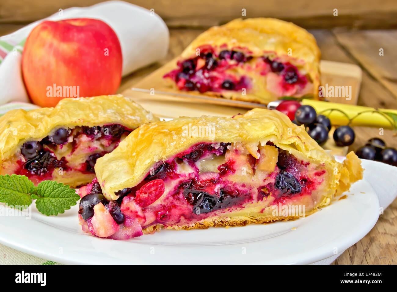 Strudel with black currant, apple and cherry on a plate, napkin, apple on the background of wooden boards Stock Photo