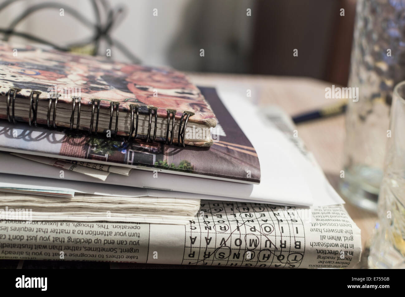A stack of magazines, newspapers and books on a table next to drinking glasses. Completed crossword visible upside down. Stock Photo