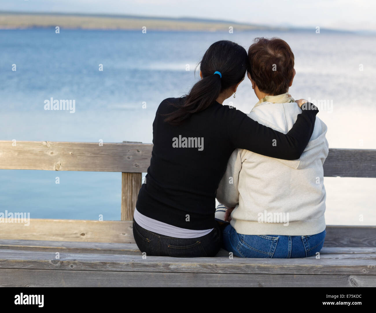 Closeup of mother with mature daughter holding her while sitting on wooden bench looking outward at the lake Stock Photo