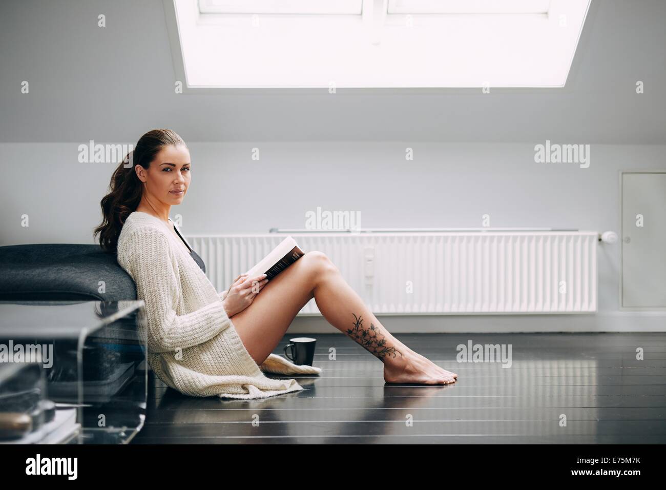 Beautiful brunette woman reading a book by a sofa. Attractive female model sitting on floor with a book looking at camera. Stock Photo