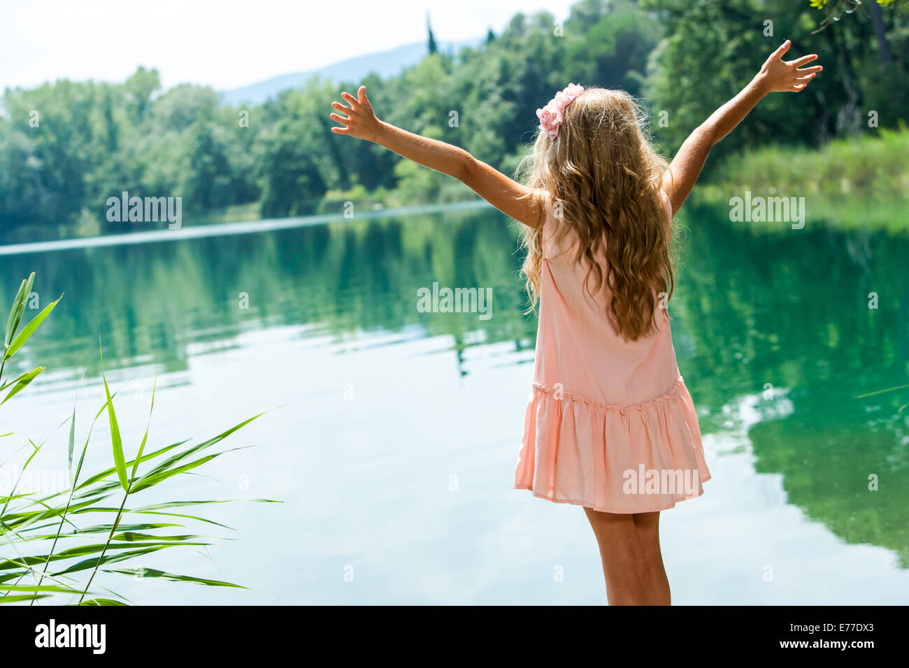 Young girl in pink dress standing with open arms at lakeside. Stock Photo